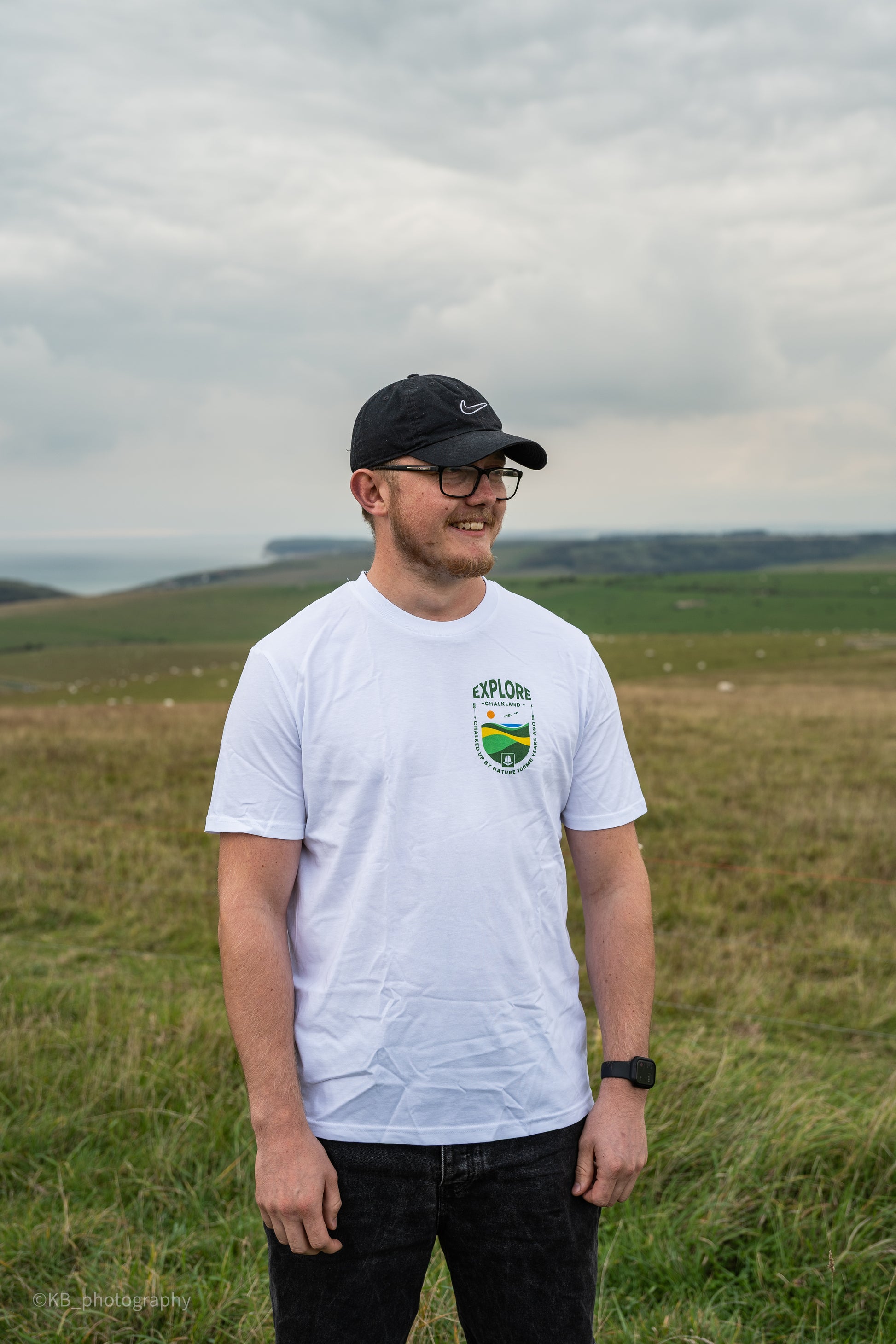 Man wearing a white t-shirt with a logo, black cap, and glasses standing in a grassy field with a cloudy sky.
