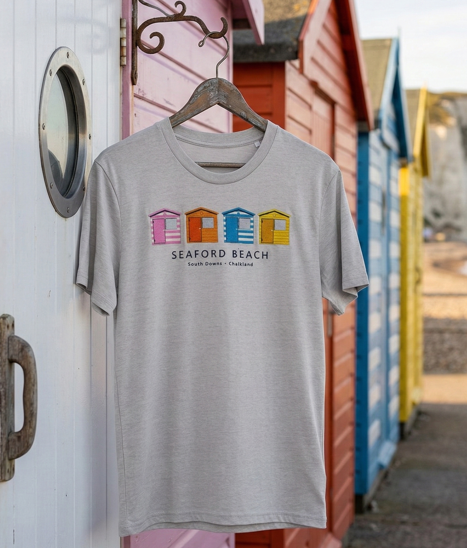 Gray t-shirt with colorful text design hanging on a wooden post with beach huts in the background