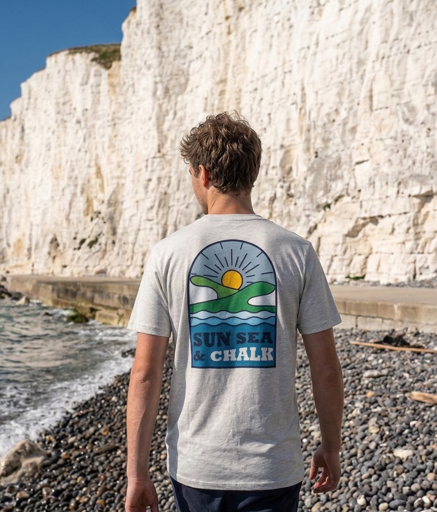 Person wearing a t-shirt with 'Sun Sea Chalk' design on a beach with white cliffs.