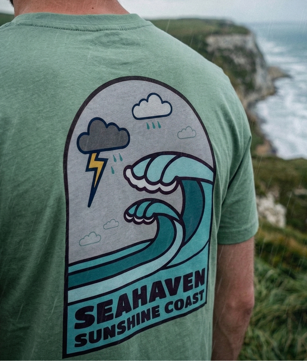 Person wearing a green t-shirt with a wave design and 'Seahaven Sunshine Coast' text, standing on a cliff overlooking the ocean.